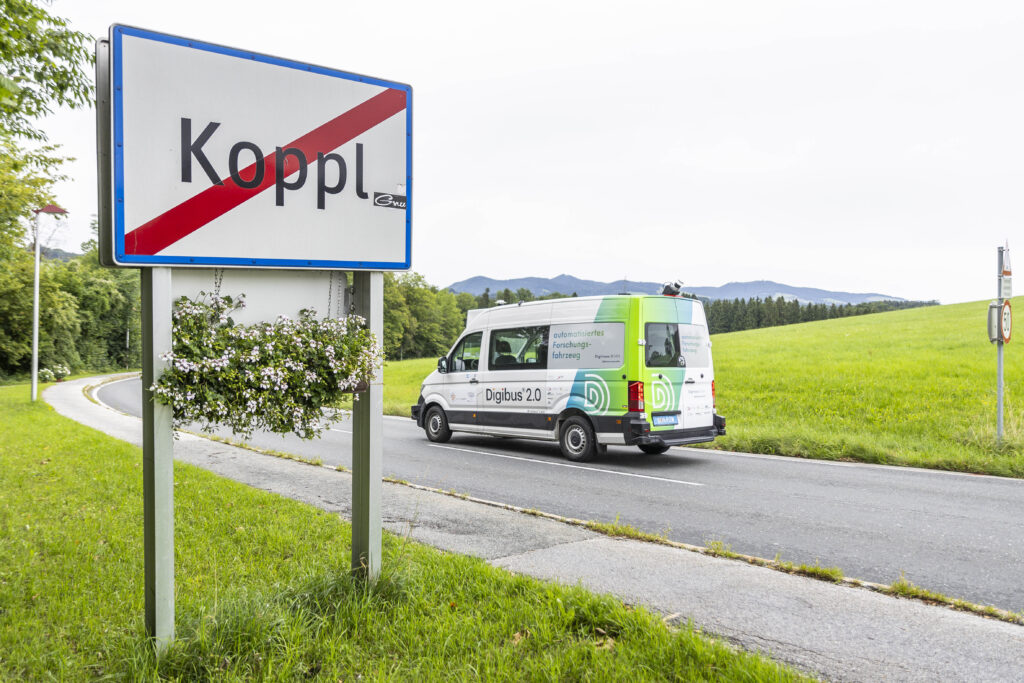 Ortsausgangsschild von Koppl mit vorbeifahrendem Digibus